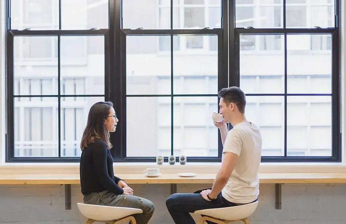 Two people talking over coffee at a long counter in front of large windows.